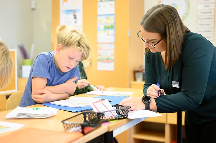 En pojke sitter vid en bänk i ett klassrum och läser medan en lärare står mittemot eleven och lyssnar på eleven.