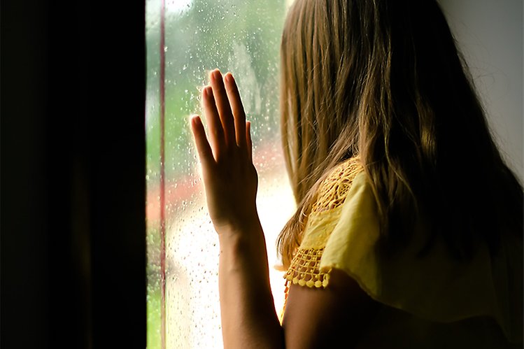 Bilden visar en flicka som står vid ett fönster och tittar ut. 