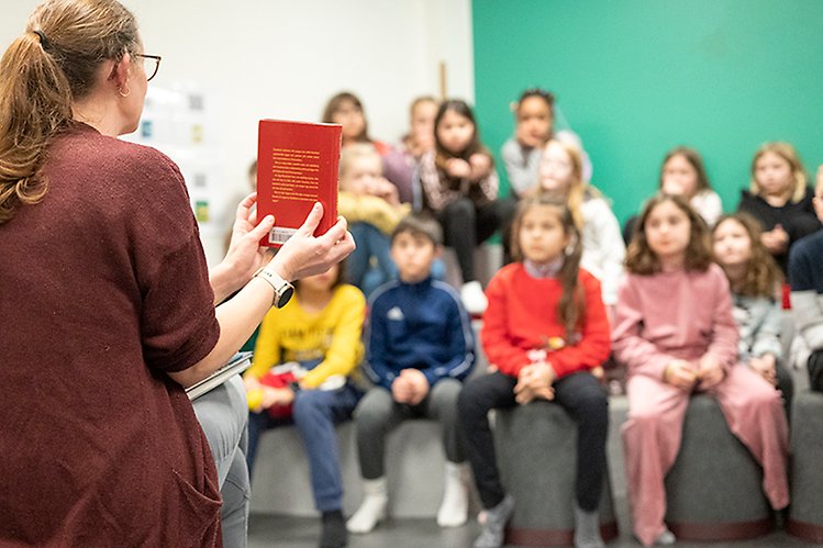 Förskollärare håller upp röd bok inför en grupp förskolebarn i bakgrunden